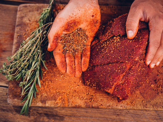 Hands applying smoked meat seasoning to a large cut of meat before smoking